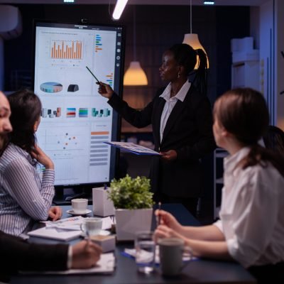 Workaholic african american leader standing in front of presentation monitor explaining marketing project solution late at night in company meeting room. Diverse teamwork working at business ideas.